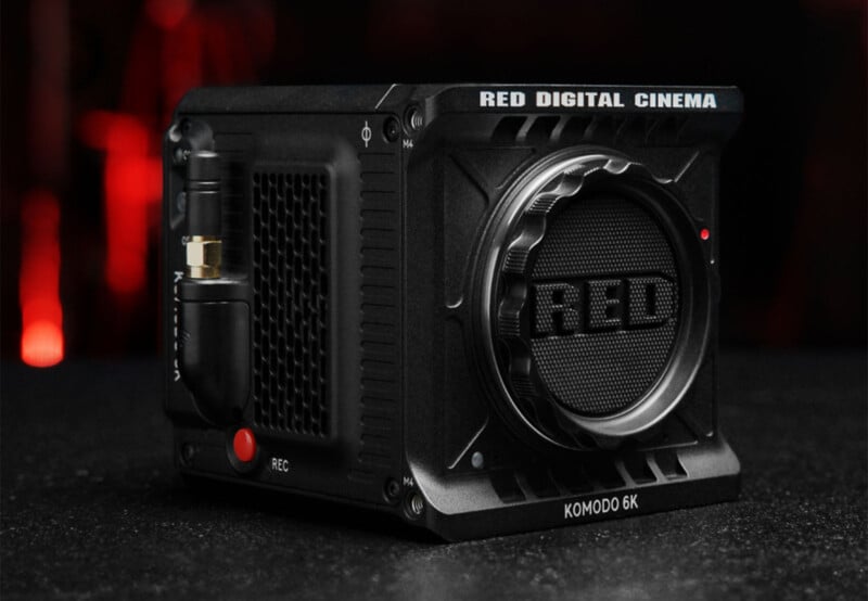 Compact black RED Digital Cinema Komodo 6K camera with textured surface, large lens mount, and visible ports, sitting on a dark surface. The camera features a prominent "RED" logo and has red and black accents. Background is dark with a blurred hint of red lighting.