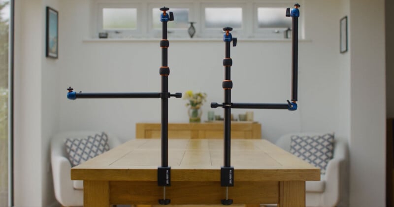 Two camera mounts are clamped to the edge of a wooden table. Each mount has adjustable sections with blue knobs and orange accents. In the background, there are armchairs with patterned cushions and a side table, as well as three small windows on the back wall.