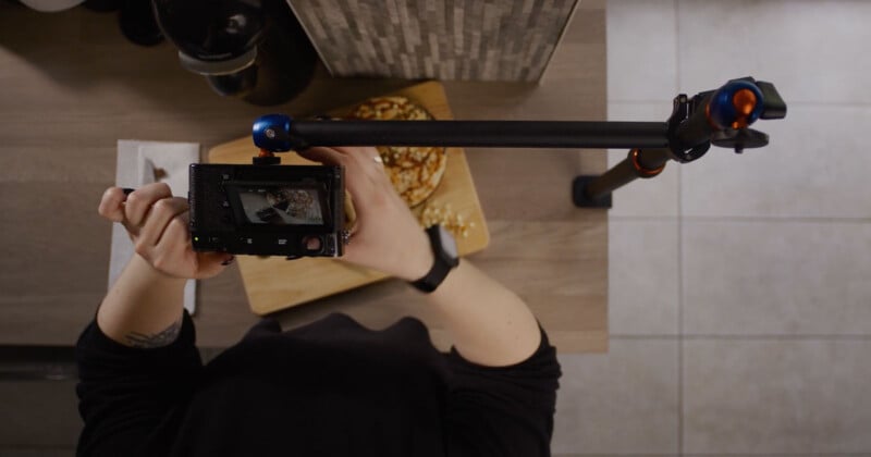 Overhead view of a person holding a camera on a boom arm, aimed at a wooden cutting board with food on it. The person is wearing a black long-sleeved shirt and a wristwatch, focusing on capturing the food from above. The background features a tiled floor and another counter.