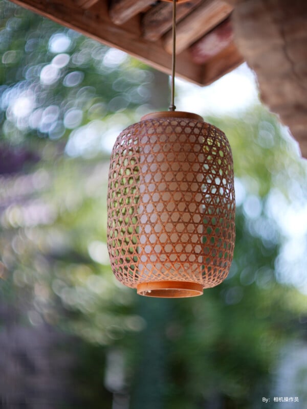 Close-up of a hanging woven lantern under a wooden roof. The lantern is cylindrical, made of bamboo or wicker, and features an open lattice design. The background is blurred with soft green and white bokeh from foliage and sunlight.