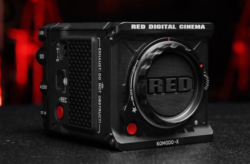 A black RED Digital Cinema Komodo-X camera is shown on a dark surface with a blurred red background. The camera features a large focus ring, various ports including USB and SDI, and several buttons including a prominent red record button.