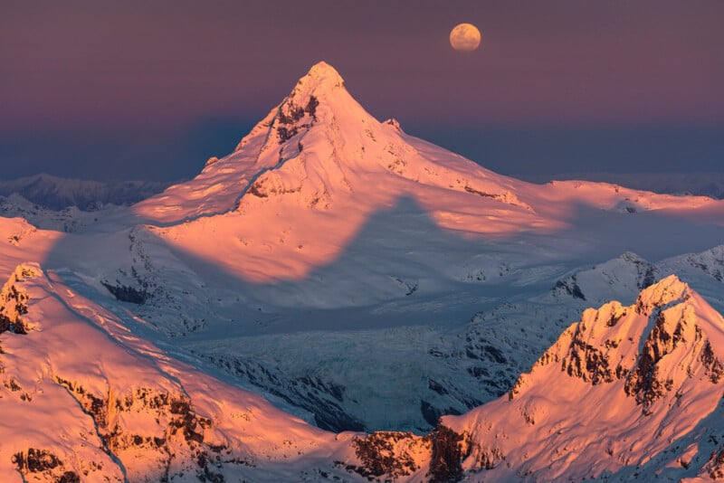 A rugged mountain peak covered in snow and bathed in a warm, pinkish-orange light, stands prominently under a clear sky. The moon is visible in the background. Surrounding the peak are snow-covered ridges and valleys, casting long shadows on the snowy landscape.