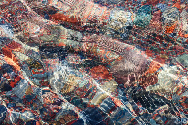 A vibrant underwater scene shows multicolored rocks and pebbles at the bottom of a clear, shallow stream. The sunlight shining through the water creates shimmering, wavy patterns on the stones.