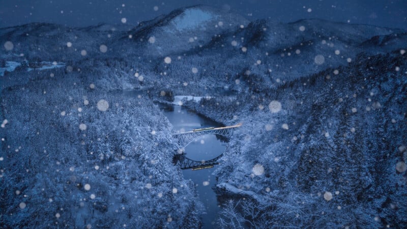 A serene winter scene depicts a snowy landscape with a railway bridge crossing over a river, surrounded by snow-covered trees. Snowflakes gently fall from the sky, adding to the tranquil atmosphere. A mountainous backdrop completes the picturesque view.