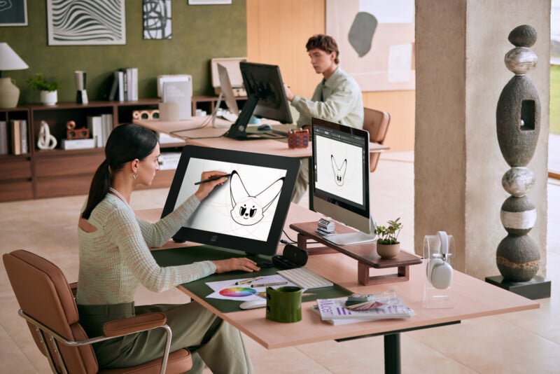 Two people are working at desks in a modern, well-lit office. The person in the foreground uses a digital tablet to draw a cat. The other person in the background is focused on their computer. The office décor includes shelves with plants and art pieces, and a sculptural column.