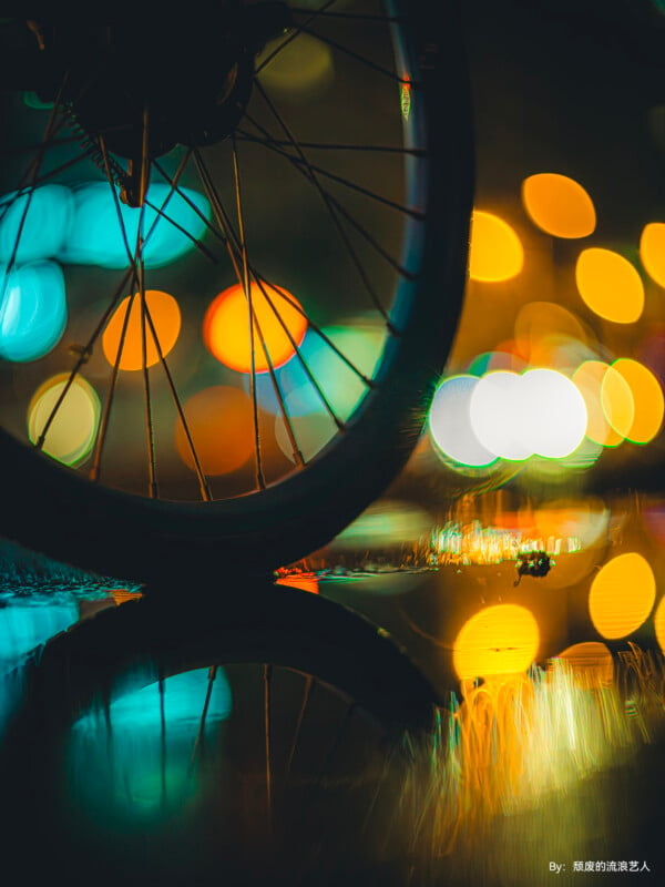 A bicycle wheel is viewed from the side, reflecting in a puddle. The background is filled with bokeh lights in various colors, creating a vibrant and abstract effect. The colors and reflections make the scene visually striking and artistic.