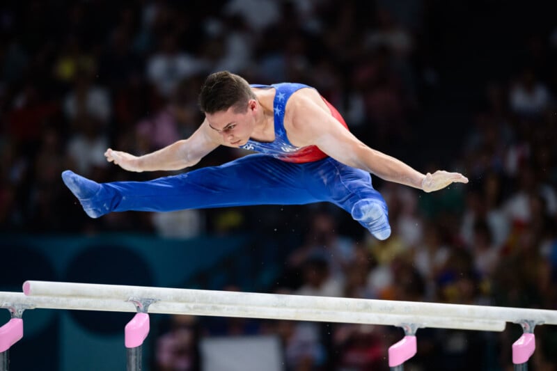 A male gymnast in mid-air performs a split above the parallel bars, dressed in a blue and red leotard, with a large audience blurred in the background.