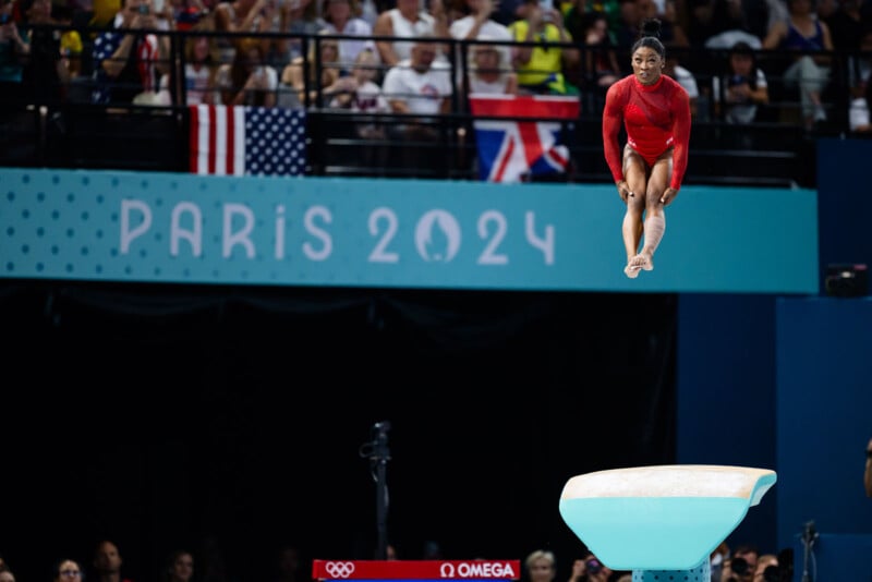 A gymnast in a red leotard is airborne, performing a vault routine at the Paris 2024 Olympics. The audience watches in the background, with flags of various countries visible. The sign behind reads "Paris 2024".