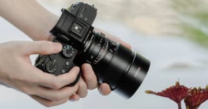 A person holding a black Olympus camera and focusing on a red leaf. The camera's dials and zoom lens are clearly visible. The background is blurred, emphasizing the camera and the leaf in the foreground.