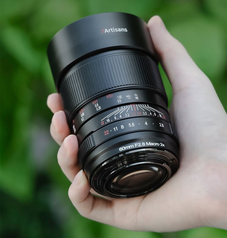 A hand holding a 7Artisans 60mm F2.8 Macro 2x camera lens, with green foliage in the background. The lens is black with red and white markings on it, showing the focus distances and aperture settings.