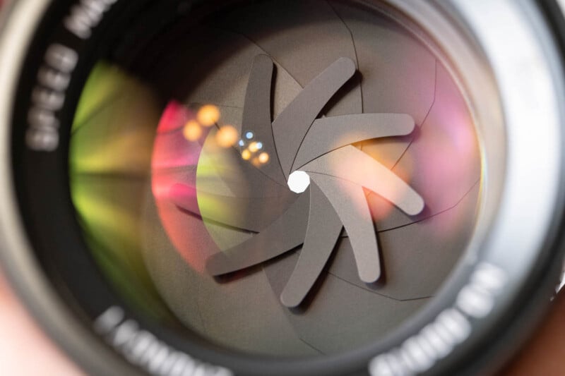 Close-up view of a camera lens aperture with partially closed blades, reflecting light in various hues of green, pink, and yellow.