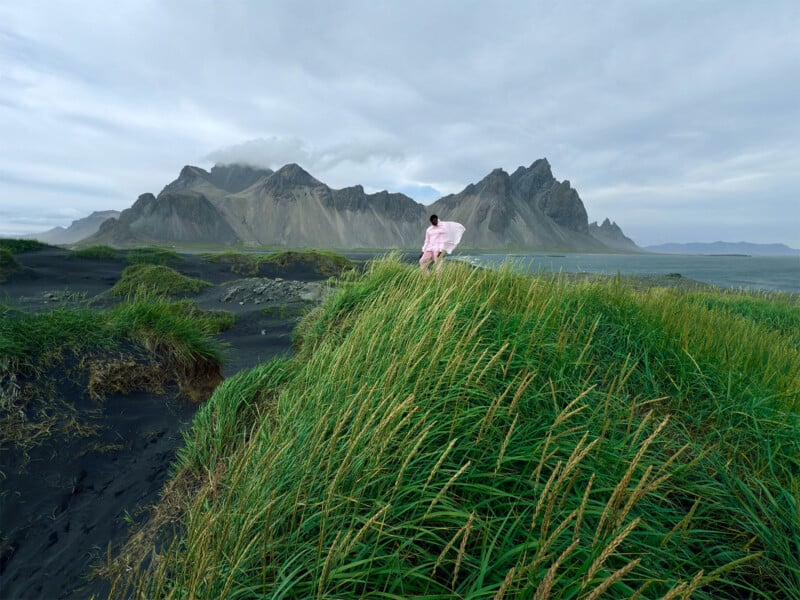 A person in a pink dress stands atop a grassy hill with dramatic, rugged mountains and a cloudy sky in the background. The landscape is a mix of green grass and dark sand, creating a striking contrast.