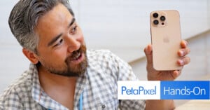 A man with gray hair, beard, and a checkered shirt holds up a gold iPhone with a triple camera setup, looking at it with interest. The image includes the "PetaPixel Hands-On" logo at the bottom right.