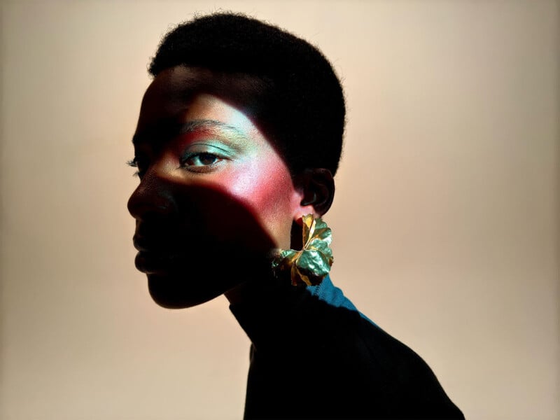 A person with short hair poses against a neutral background. They have vibrant, colorful makeup, with shades of green, pink, and blue on their face. They are wearing a large, eye-catching earring shaped like a leaf. Light and shadow dramatically highlight their features.