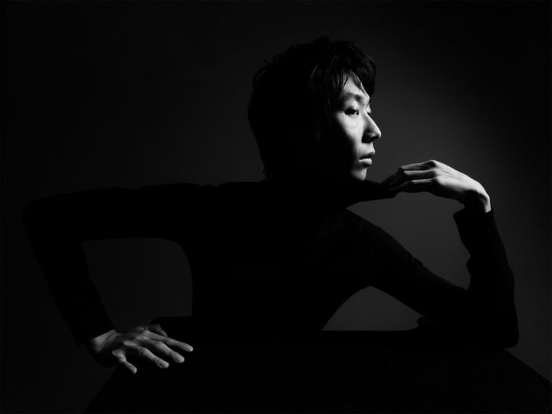 Black and white portrait of a person sitting with one arm resting on a table and the other hand gently touching their chin. They are looking to the side, illuminated from the front right, creating dramatic shadows. The background is dark.