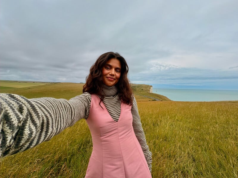 A person with long dark hair taking a selfie in a grassy field near the coast. They are wearing a striped long-sleeve shirt under a pink sleeveless dress. The sky is overcast, and a cliff and ocean are visible in the background.