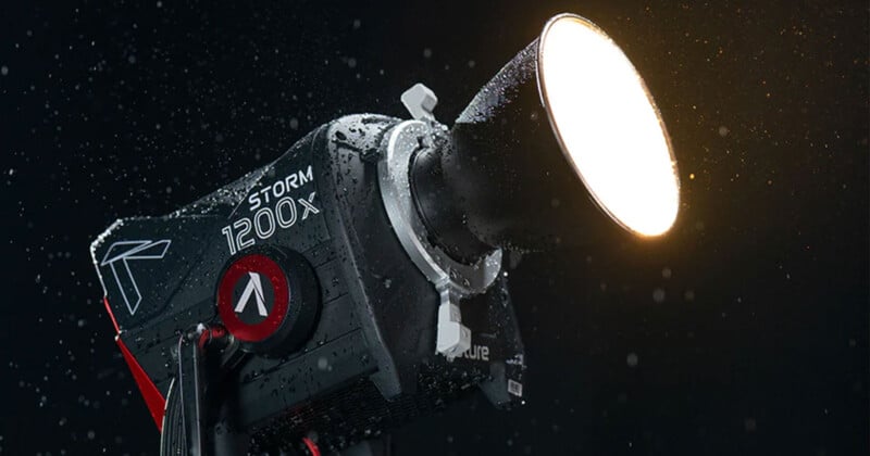 A powerful studio light, labeled "Aputure STORM 1200X," is illuminated and shown in a close-up shot against a dark background. The light is wet, covered with droplets of water, suggesting it is weather-resistant or in use during rain.