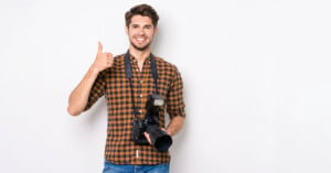 A man with short dark hair and a beard, wearing a brown plaid shirt and blue jeans, is smiling at the camera. He is holding a DSLR camera with a flash attached and giving a thumbs-up with his other hand. The background is plain white.