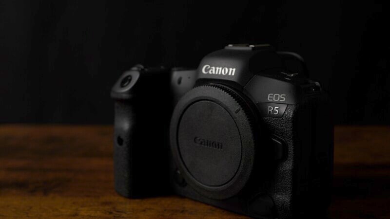 A Canon EOS R5 camera is placed on a wooden surface against a dark background. The camera is slightly angled to the left, showcasing its brand and model prominently on the front. The lens mount cover is attached, and the overall lighting is low.