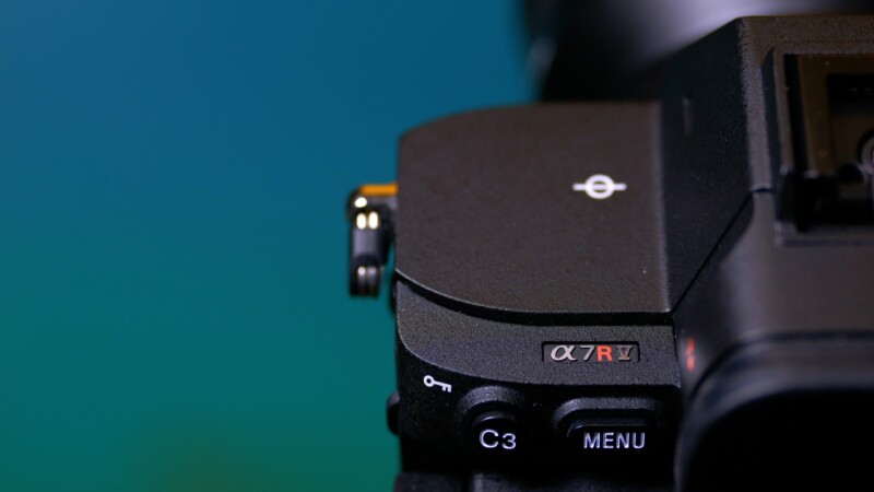 A close-up shot of a camera's top right section featuring a control dial and buttons labeled “C3” and “MENU.” The model identifier “α7R V” is visible on the camera body. The background is a gradient of teal and green.