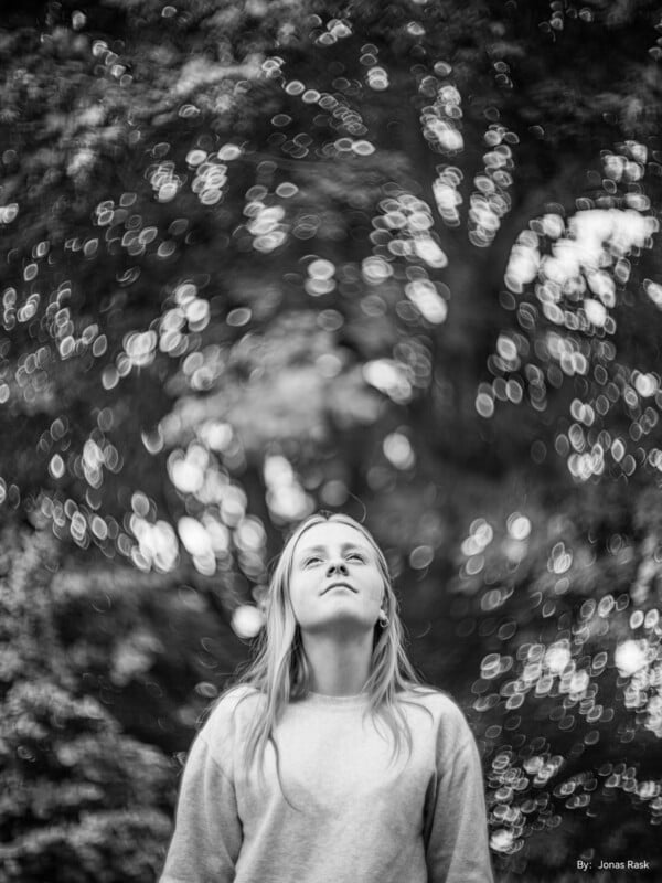 Black and white photo of a woman with long hair, looking up thoughtfully. She is wearing a light-colored sweater, standing against a backdrop of trees with a bokeh effect. The image is credited to Jonas Rask.