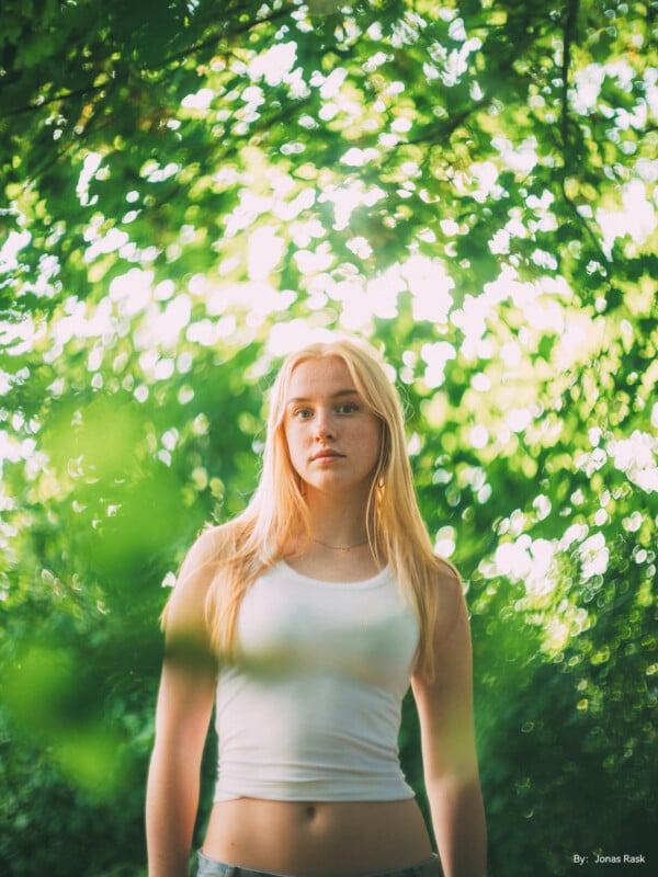 A young woman with long blonde hair stands outdoors, surrounded by lush green foliage. She is wearing a white tank top and looks ahead with a neutral expression. The sunlight filters through the leaves, creating a soft, bokeh effect in the background. By: Jonas Rask.