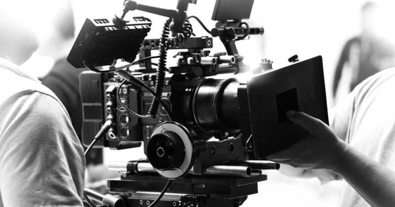 A black-and-white image of a professional film camera setup. The camera is mounted on a tripod, surrounded by various technical components and accessories. Two people are partially visible, one adjusting the camera’s lens and the other in the background.