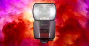 A Canon camera flash with a black body and large rectangular flash panel is centered against a vibrant, swirling background of purples, reds, and oranges.