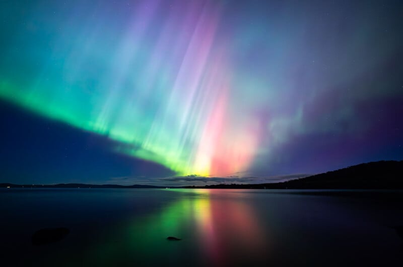 A serene night sky illuminated by aurora borealis, showcasing vibrant hues of green, purple, and pink. The colorful lights reflect beautifully on a calm body of water below. Silhouettes of distant landforms are visible under the mesmerizing display.