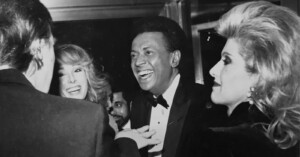 A black-and-white photo depicts a group of people, three women and one man, in a lively and happy interaction. The man and one of the women are dressed formally, possibly at a social event, and are smiling brightly as they engage in conversation.