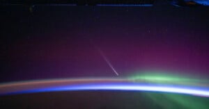 A stunning view from space captures a comet with a bright tail streaking through a star-filled sky. Below, an ethereal display of auroras in green and purple hues arcs over the curved horizon of Earth, where the atmosphere glows with a gradient of blue and orange.