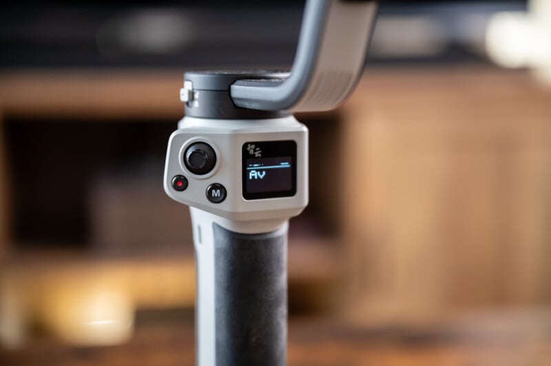 A close-up shot of a handheld gimbal stabilizer for a camera. The device has a small digital display screen showing "AV" and several buttons for controls. The background is slightly blurred, revealing indoor wooden furniture.