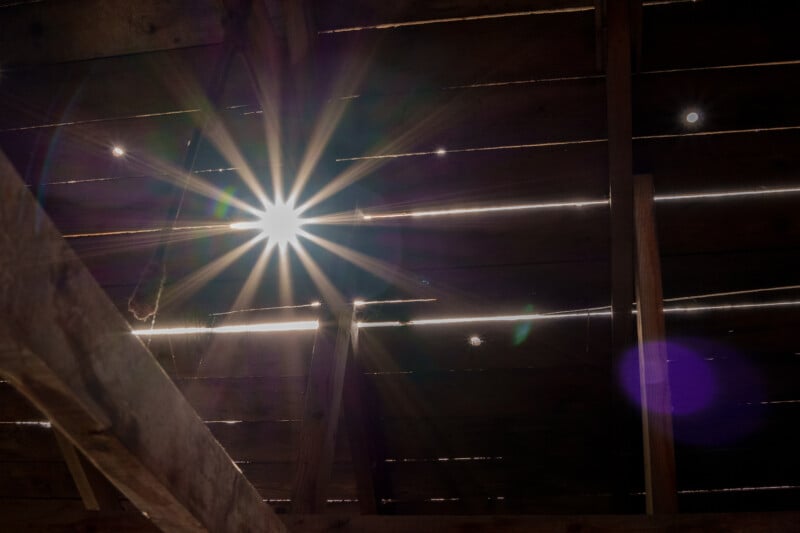 Sunlight streams through the wooden slats of a rustic roof, creating rays of light and lens flares. The light highlights the texture of the wood and casts subtle shadows, creating a warm, ethereal atmosphere.