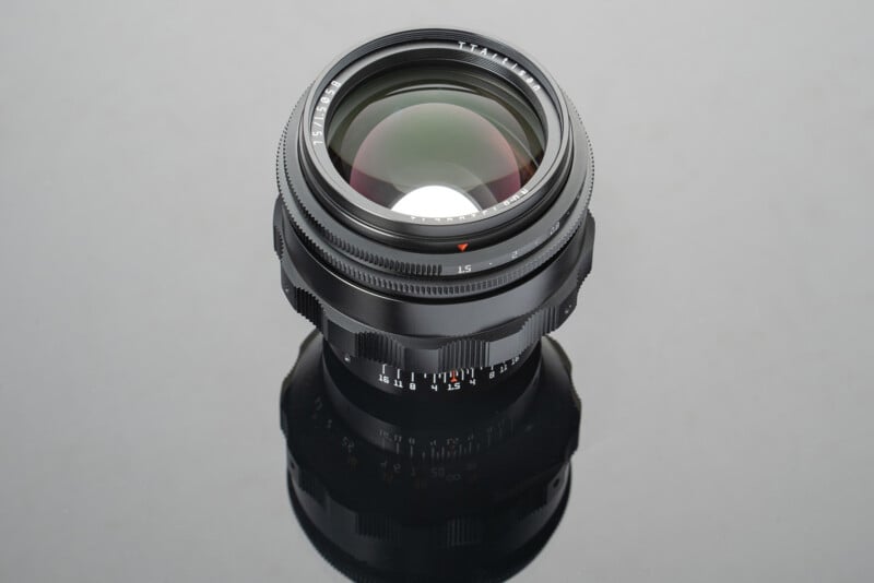 A black camera lens with various adjustment rings and markings is placed against a reflective gray surface. The lens features a red alignment dot and white labeling for focal lengths and aperture settings. The reflection of the lens is visible below it.