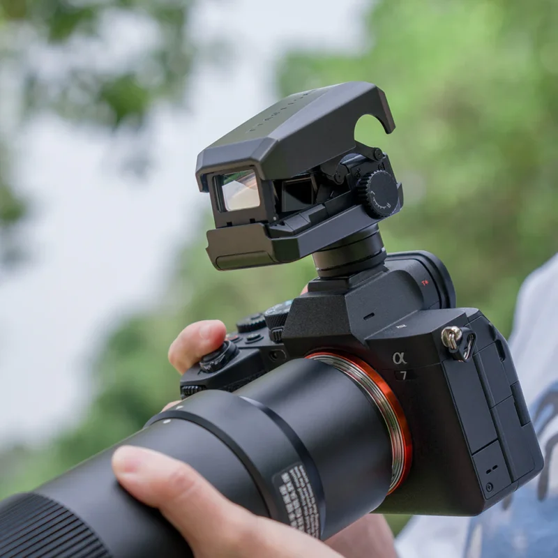 A person holds a black Sony Alpha 7 camera equipped with a large telephoto lens and a viewfinder accessory on top. The background is outdoors with blurred greenery, suggesting the person is possibly engaged in nature or wildlife photography.