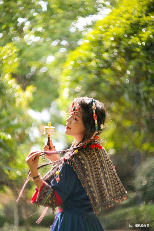 A person dressed in traditional attire stands in a sunlit, green forest. They hold a ceremonial object and wear ornate headgear adorned with colorful beads and flowers. The background is a blur of vibrant greens, giving a dreamy effect to the scene.