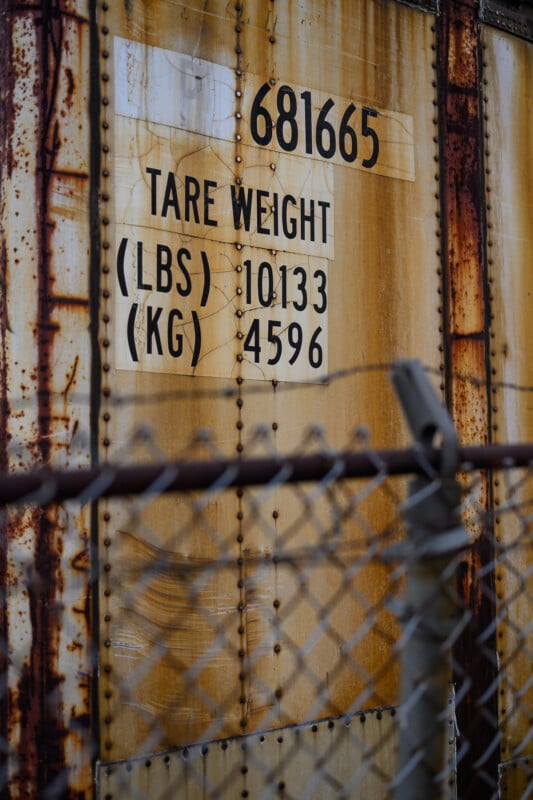 A weathered, rust-stained metal surface displays the following text: "681665, Tare Weight, (LBS) 10133, (KG) 4596". In front of the surface is a chain-link fence slightly out of focus.