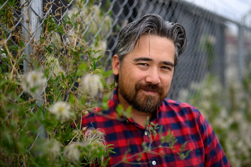 A man with salt-and-pepper hair and a full beard, wearing a red and black plaid shirt, leans against a fence entwined with green vines and small flowers. He is smiling gently, and the background is slightly blurred.