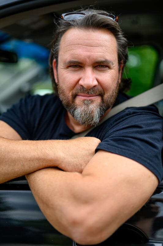 A middle-aged man with a beard and mustache is leaning out of a car window, smiling. He is wearing a black t-shirt and has sunglasses perched on his head. His arms are crossed on the car door, and a seatbelt is visible. The background is slightly blurred with greenery.