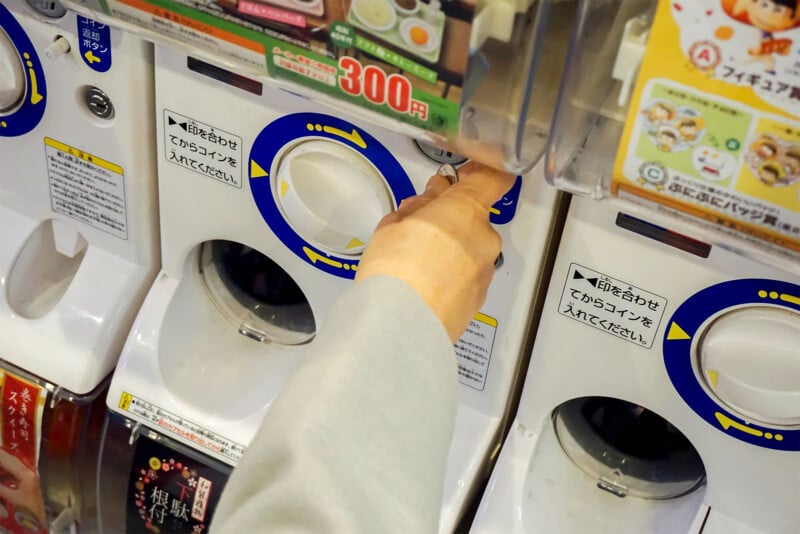 A person inserts coins into a capsule toy vending machine, known as a gashapon, in a brightly lit area. The machine displays toy options and prices. The person's hand is turning the knob to dispense a toy capsule.