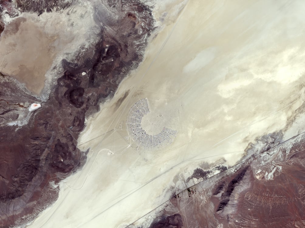 Aerial view of a semi-circular desert settlement in the center of a vast, dry landscape. Surrounding the settlement are rocky, barren terrains of varying shades. The settlement consists of numerous small structures arranged in an organized, radial pattern.