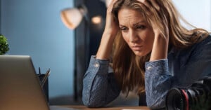 A woman with long blond hair sits at a desk, looking stressed. She holds her head with both hands while staring at an open laptop screen. A camera lens and a plant are visible in the background. The setting appears to be an office or a workspace.