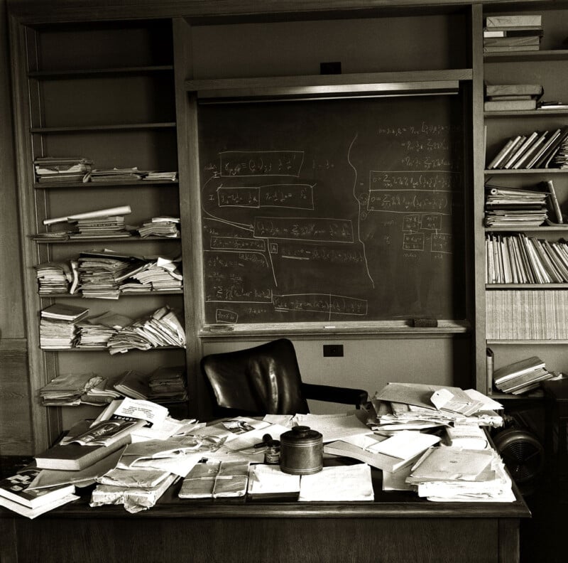 A black and white photo of a cluttered office featuring a desk covered in papers, books, and a coffee pot. Behind the desk is a chalkboard filled with hand-written mathematical equations. Books and stacks of documents fill the shelves on either side of the chalkboard.