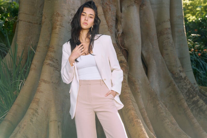 A woman with long dark hair stands pensively in front of a large tree trunk. She is dressed in a light beige blazer over a white top and matching beige pants, with one hand in her pocket and the other touching her chest, looking off into the distance.