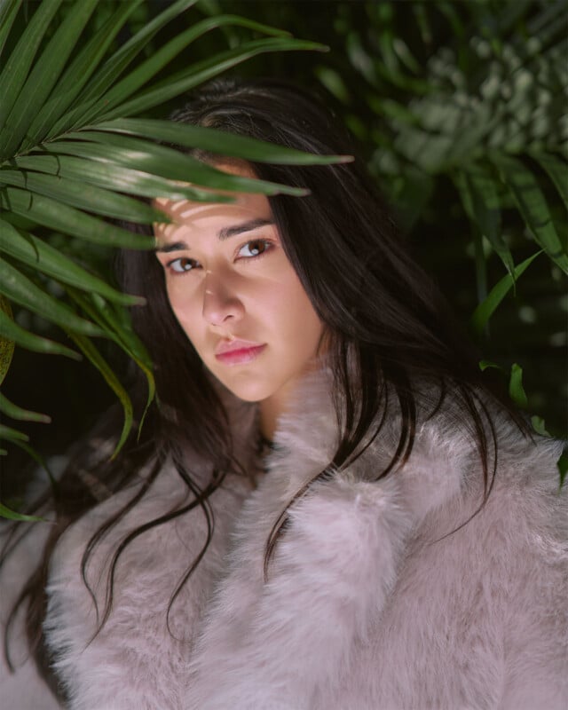 A woman with long dark hair, wearing a fluffy light-colored coat, gazes into the camera with a serious expression. She is partially hidden behind large green palm leaves, creating a contrast between the vibrant foliage and her light coat.