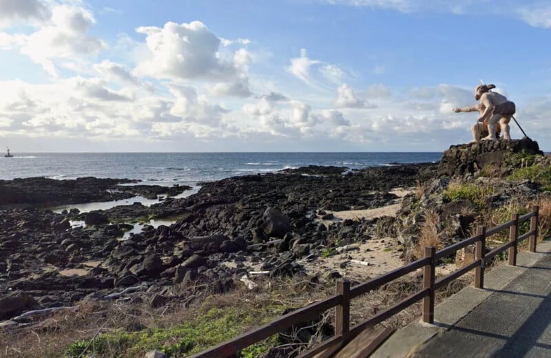 A coastal scene with rocky shorelines and clear skies. A large, white, statue of a cow and person stands on an elevated rock formation to the right, facing the ocean. A wooden fence lines a pathway in the bottom right corner of the image.