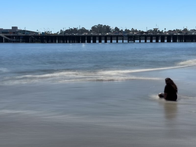 A person sits by the water’s edge on a sandy beach, facing the ocean. The scene is tranquil with gentle waves lapping at the shore. In the distance, a pier extends out into the sea, and a town with trees and buildings lines the horizon under a clear blue sky.