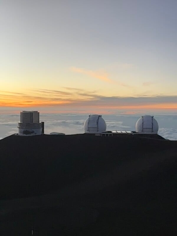 Sunset view of an observatory on a mountain peak, featuring three large dome-shaped telescopes. The sky is a blend of soft oranges and blues, with clouds floating below the observatory, giving an impression of being above the clouds.