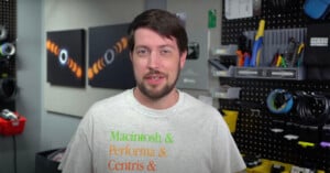 A person with short dark hair and a beard stands in a workshop-like setting. They are wearing a light gray T-shirt with green and orange text that reads "Macintosh & Performa & Centris & Quadra." Various tools, cables, and supplies are organized on shelves and pegboards behind them.