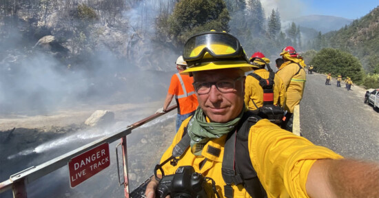 A person in yellow fire protective gear and a helmet takes a selfie with a group of firefighters in the background standing on a road near a smoky forest fire. A sign on the left reads "DANGER LIVE TRACK." Trees and hills are visible beyond the smoke.